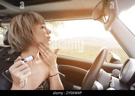 Niedliche blonde Frau mit kurzen Haaren, die Lippen färben, während sie am Steuer ihres Autos sitzt. Das Konzept, eine Frau zu fahren. Unangemessene Fahrstunden. D Stockfoto