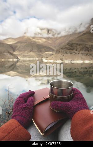 Die Frauenhände in Mänteln und Fäustlingen aus der ersten Person halten einen Metallbecher mit Tee oder Kaffee und ein Ledertaschenbuch vor dem Hintergrund Stockfoto