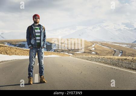 Ein stylischer bärtiger Hipster in Sonnenbrille mit einem Vintage-Rucksack steht an einem sonnigen Tag auf einem Landstraßenasphalt. Das Konzept des Anhalter- und Wanderns Stockfoto