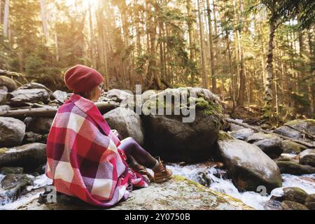 Ein hübsches Hipster-Mädchen mit einem karierten Karo auf den Schultern, einem Hut, einem gelben Pullover und einer Brille mit einer Tasse Kaffee in den Händen schaut auf eine Halterung Stockfoto