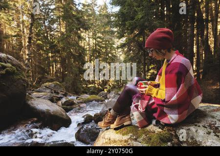 Ein hübsches Hipster-Mädchen mit einem karierten Karo auf den Schultern, einem Hut, einem gelben Pullover und einer Brille mit einer Tasse Kaffee in den Händen schaut auf eine Halterung Stockfoto