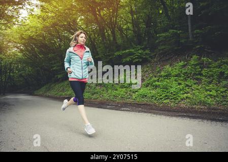 Junge Fitness blonde Frau in Kopfhörer läuft am Morgen kaukasischen Wald Trail in Sonnenlicht Stockfoto