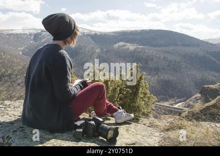 Porträt eines Hipster-Designers Fotografin mit Hut und Sonnenbrille zeichnet ein Pastellpapier in ihr Notizbuch, das draußen auf einem Felsen in den Bergen sitzt. Die Stockfoto