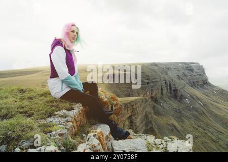 Ein Mädchen-Reisende mit bunten Haaren sitzt am Rande einer Klippe und blickt auf den Horizont auf einem Hintergrund eines felsigen Plateau Stockfoto