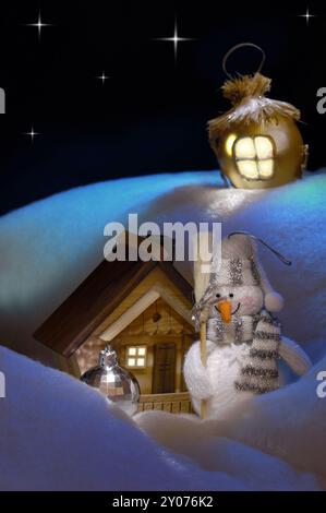 Helles konzeptionelles Weihnachtsstillleben mit einem Schneemann, der neben einem Holzhaus unter einem schneebedeckten Hügel steht. Künstlerische Weihnachtskarte, Rea Stockfoto