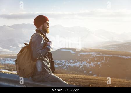 Bärtiger Touristen-Hipster-Mann in Sonnenbrille mit einem Rucksack, der auf einem Straßenrand sitzt und den Sonnenuntergang vor dem Hintergrund eines schneebedeckten M beobachtet Stockfoto