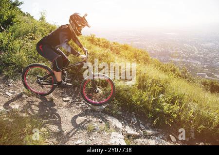 Im Sommer an einem sonnigen Tag fährt Ryder mit einem Helm vom Berg aus mit dem Mountainbike vor der Kulisse einer Stadt in der den Schotterweg hinunter Stockfoto