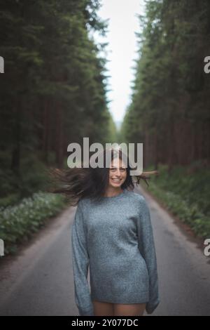 Eine junge Frau mit fließenden dunklen Haaren steht selbstbewusst auf einer Waldstraße, ihr Haar wird vom Wind gehoben. Eine Mischung aus Freiheit, Natur und jugendlicher Energie c Stockfoto