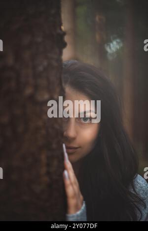 Rätselhafte Frau mit fesselnden blauen Augen blickt hinter einem Baum in einem mystischen Wald. Das Bild fängt das Wesen des Geheimnisses, der Natur und der Ruhe ein Stockfoto