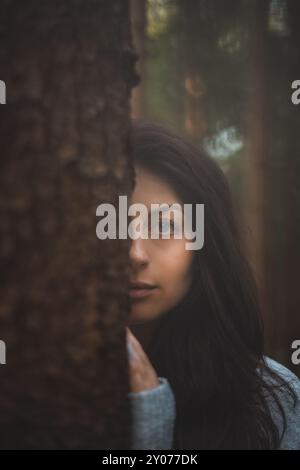 Rätselhafte Frau mit fesselnden blauen Augen blickt hinter einem Baum in einem mystischen Wald. Das Bild fängt das Wesen des Geheimnisses, der Natur und der Ruhe ein Stockfoto