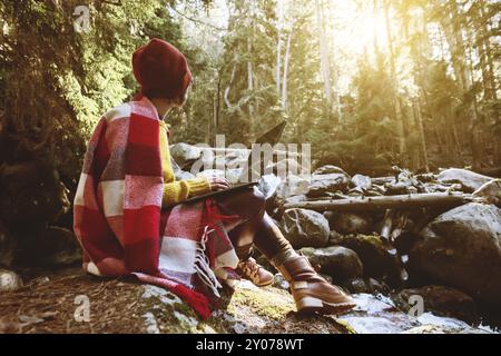 Ein farblich abgestimmtes Porträt eines freiberuflichen Hipster-Mädchens mit Brille und einer stilvollen Mütze, die in einer Decke gekleidet ist, mit einem Laptop, der kniend auf einem Felsen in einem Nadelholz sitzt Stockfoto