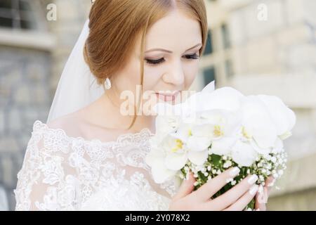 Lächelnde junge Braut in weißem Kleid schnüffelt an ihrem Hochzeitsstrauß, der sorgfältig von den Händen gehalten wird. Das Konzept der Floristen bei Hochzeiten Stockfoto