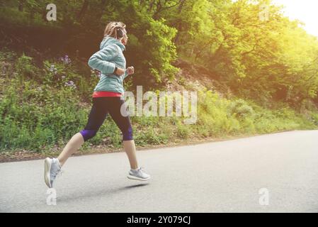 Eine junge blonde Frau, die läuft, übt im Freien in einem Bergpark der Stadt im Wald. Warme Strahlen durch die Äste der Bäume Stockfoto