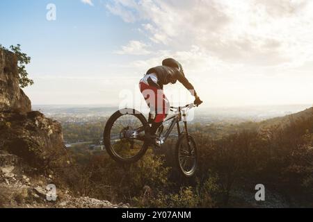 Ein junger Fahrer am Steuer seines Mountainbikes springt bei Sonnenuntergang von hohen Klippen hinunter vor dem Hintergrund der Stadtlandschaft. Bergab Bergweg in t Stockfoto