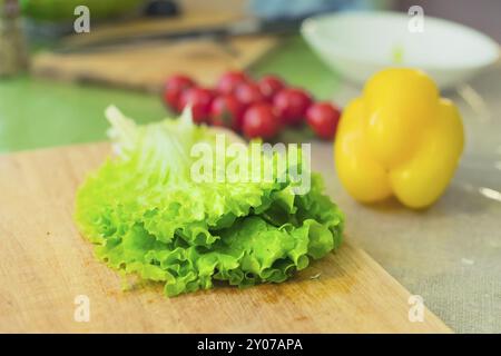 Frische grüne Salatblätter liegen auf einem hölzernen Schneidebrett neben gelbem Paprika und roten Kirschtomaten auf einem grünen Tisch. Das Konzept der gesunden Ernährung Stockfoto