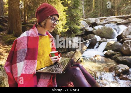 Hipster-Mädchen-Designerin mit einem Karo auf den Schultern, einem Hut und einem gelben Pullover und einer Brille mit einem Laptop auf dem Schoß und einer Tasse Kaffee in der Hand Stockfoto