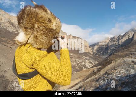 Eine Touristenfotografin mit Pelzmütze und gelbem Pullover in den Bergen macht Fotos mit ihrer Digitalkamera. Der Begriff der Fotografie auf der Reise Stockfoto