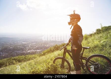 Ein junger Mann, der auf dem Mountainbike auf einem Berg steht, wenn unter den Bergen niedrige Wolken und Nebel liegen Stockfoto