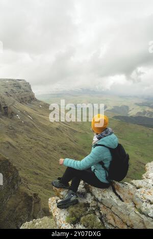 Junge Frau mit Rucksack, die pensiver am Rande eines Felsens sitzt und mit Wolken in den Himmel blickt Stockfoto