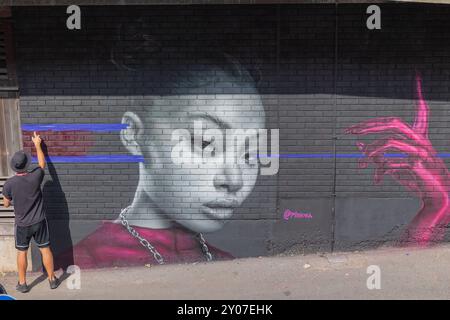 Southend on Sea, Großbritannien. Oktober 2024. Straßenkünstler arbeitet an einem großen Wandgemälde in Southend-on-Sea, Essex. Das Kunstwerk zeigt ein graues Porträt mit leuchtenden rosa und roten Akzenten, einschließlich einer Hand und Kleidung. Der Künstler bringt blaues Klebeband über die Ziegelwand an, was darauf hindeutet, dass die Arbeiten an der Gestaltung fortgeführt werden. Die Künstler arbeiten daran, ihre Arbeit am letzten Tag des Southend City Jam zu beenden. Penelope Barritt/Alamy Live News Stockfoto