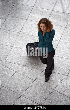 Eine junge Frau mit einem Koffer steht auf einem Platz und redet mit einem Handy Stockfoto