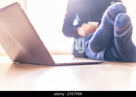 Junger Mann mit warmen Socken sitzt auf dem Holzboden und genießt den Tag, Laptop im Vordergrund. Stockfoto