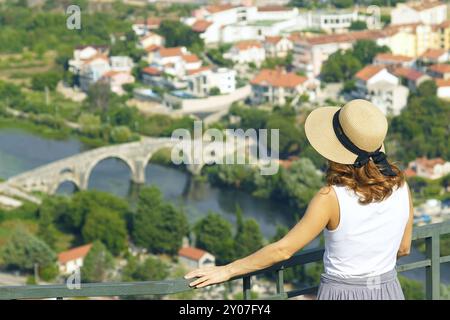 Von hinten fotografiert, bewundert eine junge Frau mit Hut den Blick von oben auf das wichtigste Wahrzeichen der Stadt Trebinje - die Arslanagische Brücke. Stockfoto