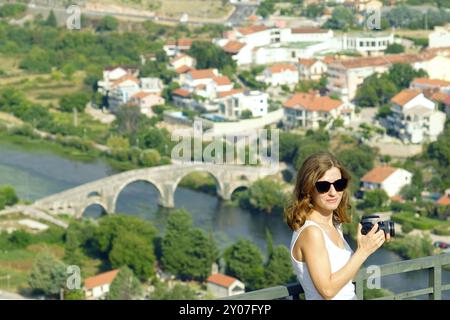 Eine attraktive Frau steht mit einer DSLR-Kamera in den Händen und ist dabei, von der Aussichtsplattform aus ein Foto von der Arslanagischen Brücke in Trebinje zu machen. Stockfoto