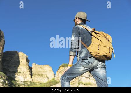 Stylischer bärtiger Reisender in Sonnenbrille und einer Mütze mit Rucksack in Denim-Anzug und gelben Schuhen steht mit dem Rücken auf einem großen Stein am Fo Stockfoto