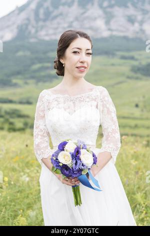 Eine wunderschöne kaukasische Braut mit einem Blumenstrauß in den Händen steht vor dem Hintergrund der Kaukasusberge und der grünen Felder Stockfoto