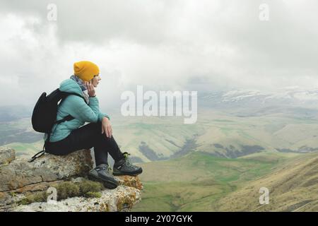 Junge Frau mit Rucksack, die pensiver am Rande eines Felsens sitzt und mit Wolken in den Himmel blickt Stockfoto