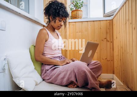 Afroamerikanische Frau in lässiger Kleidung, die ferngesteuert am Laptop arbeitet, während sie auf dem Boden in gemütlicher Wohnumgebung auf dem Balkon sitzt. Freelancer Digital Nomad arbeitet unter unangenehmen Bedingungen Stockfoto