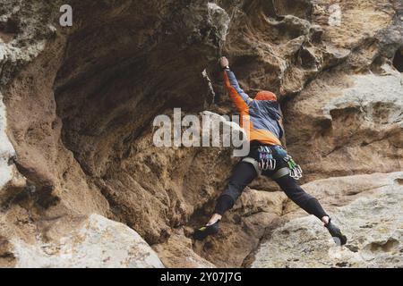 Hipster, Kletterer im Alter, in dem man einen wunderschönen Felsen ohne Versicherung und Helm hochklettert. Ein Bergsteiger in Hut und Daunenjacke mit Tasche für magnes Stockfoto