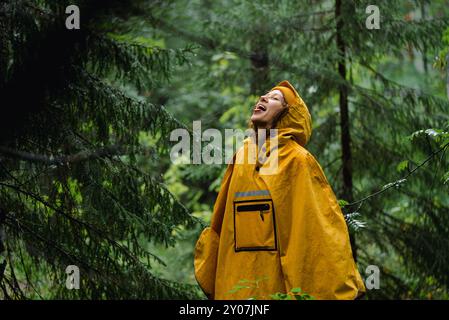 Eine Frau im Regenmantel hebt den Kopf hoch und ragt in dichtem feuerem Wald aus. Touristen in Poncho genießen bewölktes Wetter und Gewitter am Sommertag Stockfoto