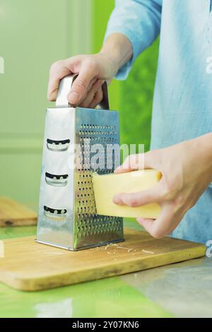 Nahaufnahme der Hände einer Frau reiben einen Parmesankäse auf einer Metallreibe auf einem grünen Tisch Stockfoto