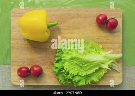 Frische grüne Salatblätter liegen auf einem hölzernen Schneidebrett neben gelbem Paprika und roten Kirschtomaten auf einem grünen Tisch. Das Konzept des gesunden f Stockfoto