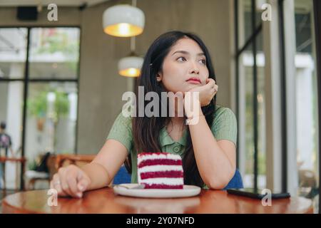 Eine Frau sitzt an einem Tisch mit einem Stück Kuchen vor sich. Sie sieht traurig aus und hält ihre Hand an ihr Gesicht. Stockfoto