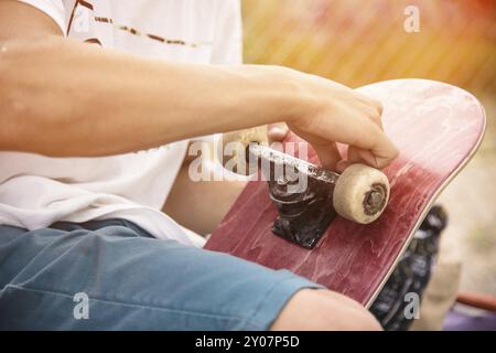 Ein kleiner Junge auf den Knien klebt das Griptape in Begleitung von Freunden bei sonnigem Wetter auf ein Skateboard. Vorbereitung eines Skateboards für einen Skatepark-Wettkampf Stockfoto