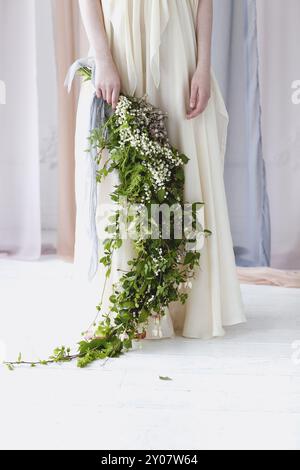 Barfuß-Frau in elegantem weißen Hochzeitskleid mit großem Blumenstrauß aus kleinen weißen Blumen und grünem Laub, das unter transparenten Pastellfarben steht c Stockfoto