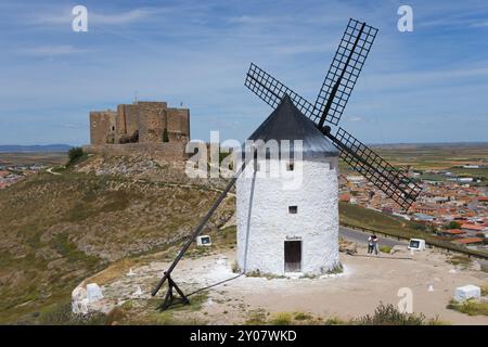 Eine alte Windmühle vor einer mittelalterlichen Burg auf einem Hügel mit einem Dorf im Hintergrund unter blauem Himmel, Consuegra, Toledo, Castilla-La Mancha, Rout Stockfoto