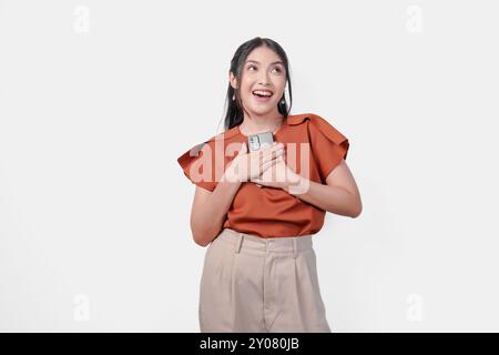Glückliche achtsame, dankbare junge asiatische Frau lächelt, während sie das Telefon mit Händen auf der Brust über isoliertem weißen Hintergrund hält. Stockfoto
