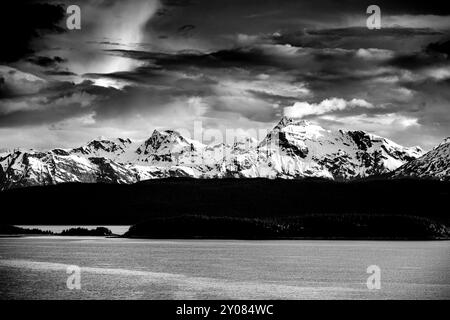 Das Meer mit einer Silhouette von Bäumen vor schneebedeckten Bergketten und dramatischen Himmeln. Alaska, usa, Stockfoto