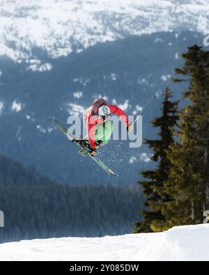 Skifahrer, der einen Sprung auf einem schneebedeckten Berg mit Kiefern und blauem Himmel im Hintergrund macht Stockfoto