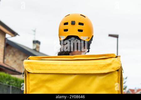 Nahaufnahme, Rückansicht eines Latina Food Delivery Girl mit gelbem Rucksack und Helm. Das Bild konzentriert sich auf die leuchtend gelbe Ausrüstung, während sie auf ihr reitet Stockfoto