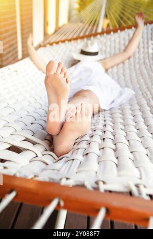 Junge Frau in weißem Kleid und Hut in Hängematte, während sie auf der Terrasse des Hauses entspannen Stockfoto