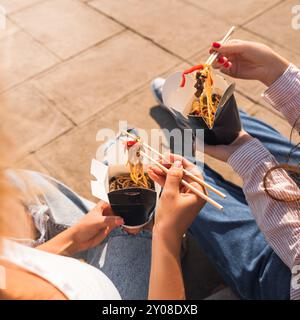 Junge Mädchen, Freundinnen essen asiatisches Essen mit Wok-Nudeln, mit Essstäbchen, Essen zum Mitnehmen, Pappschachtel mit Nudeln zum Mitnehmen Stockfoto