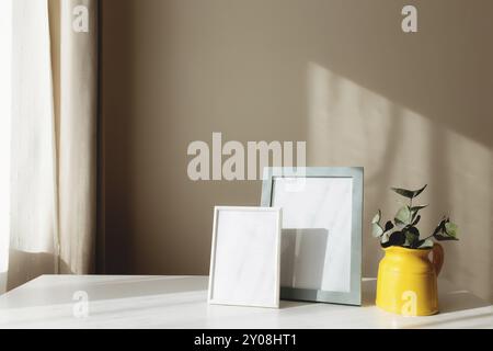 Eine gelbe Keramikkkanne oder -Vase mit Eukalyptuszweigen, leere weiße Bilderrahmen auf dem weißen Tisch im Innenraum mit beigefarbenen Wänden in der Nähe des Fensters Stockfoto