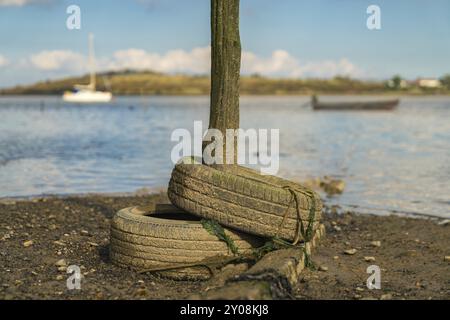 Alte Reifen und ein Holzpfosten in den Oare Marshes in der Nähe von Faversham, Kent, England, Großbritannien, mit Booten und der Isle of Sheppey im Hintergrund Stockfoto