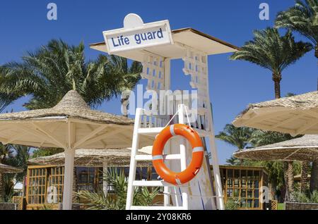 Hölzerner Rettungsschirm mit orangefarbenem Rettungsschirm am Strand vor dem Hintergrund von Palmen und Schilfschirmen Stockfoto