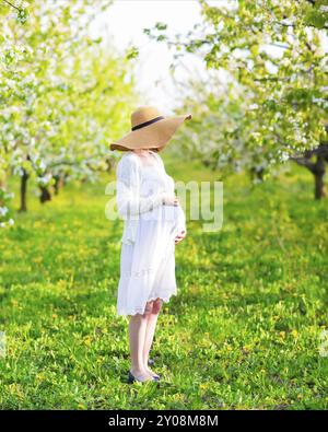 Schöne schwangere Frau mit großen Hut und weißen Kleid im blühenden Garten Stockfoto